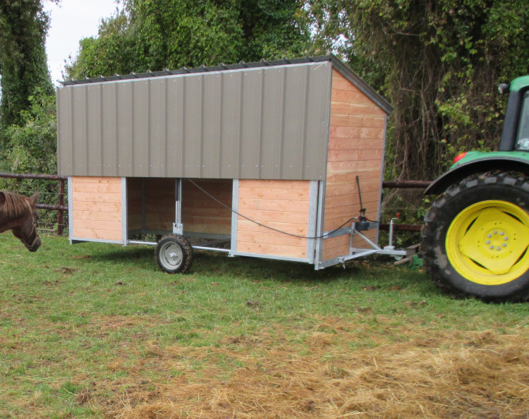 Le modèle d'abri mobile pour chevaux Extensible arrive dans la prairie attelé à un tracteur agricole surveillé par un cheval.